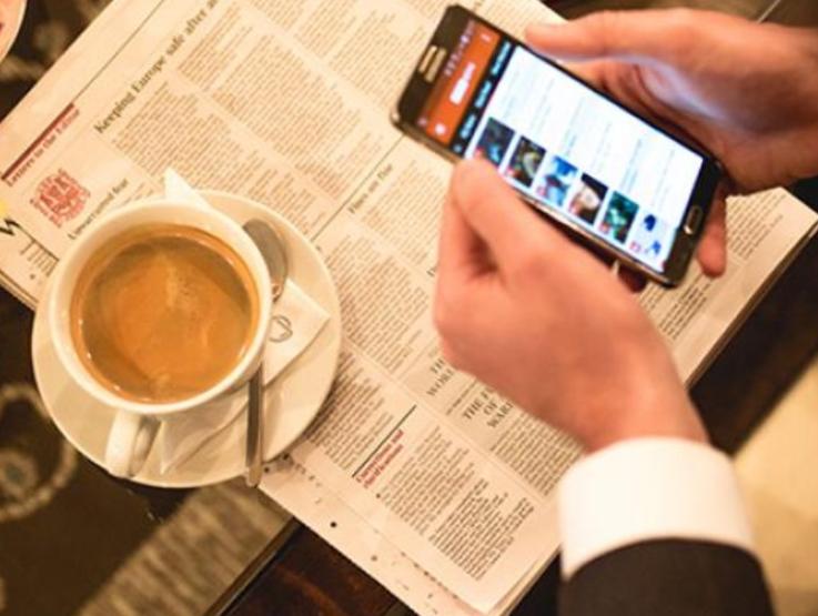 man holding phone with newspaper on table and coffee